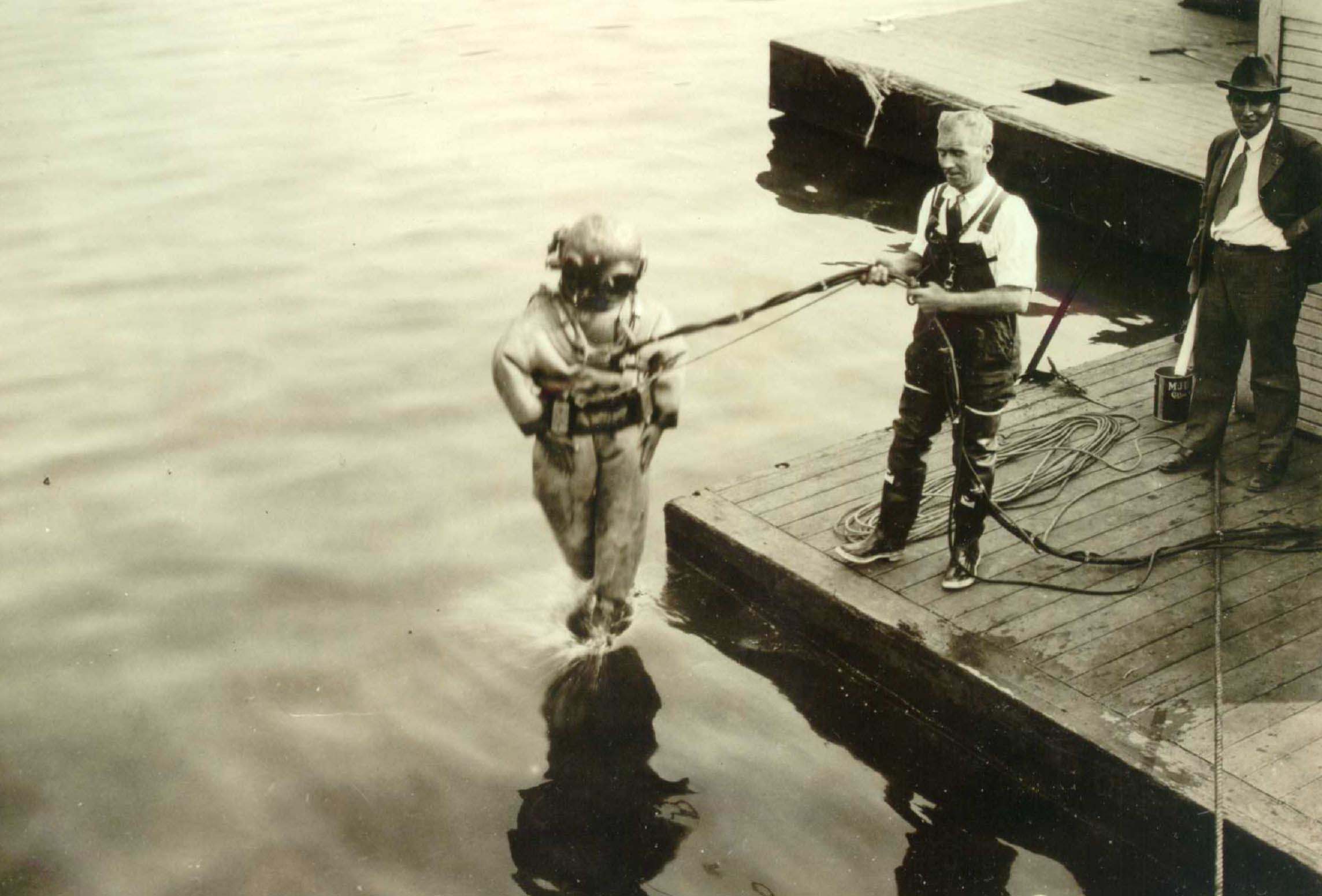 Two men standing on a dock while another in a diving suit is plunging feet first into the water. Once on the men his holding tubes and cables attached to the diving suit.